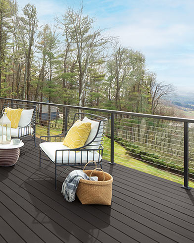 Bonitos muebles de exterior acolchados de color blanco con almohadones amarillos y una mesa pequeña en una terraza teñida con Woodluxe® liso en color Marrón Cordobán ES-62.