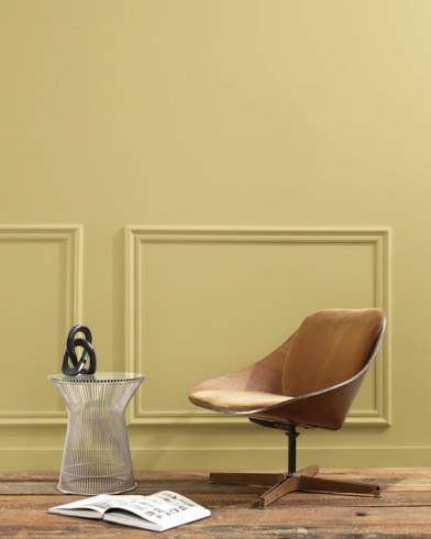 A flat wood riser holds a curved wooden chair, glass vase and open book in front of a paneled wall painted Anjou Pear.
