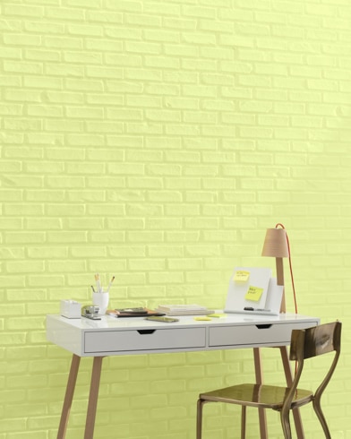 A white and copper writing desk and a bronze metal chair sit in front of a brick wall painted Apples and Pears.