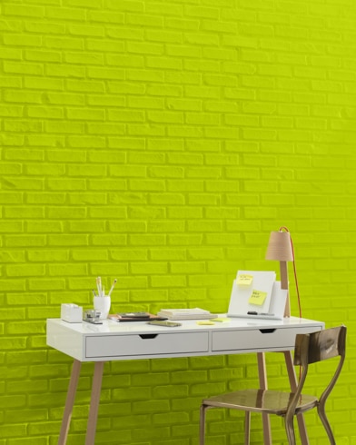 A white and copper writing desk and a bronze metal chair sit in front of a brick wall painted Bright Lime.