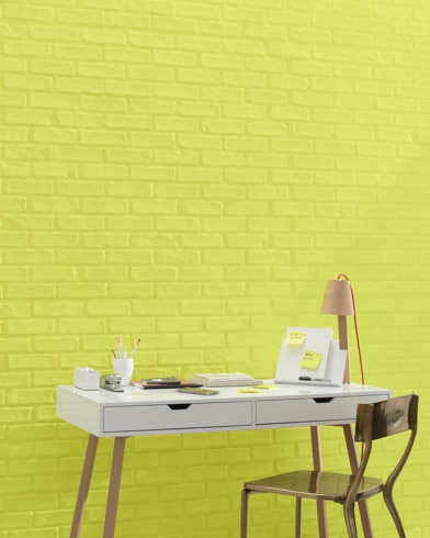 A white and copper writing desk and a bronze metal chair sit in front of a brick wall painted Chamomile.