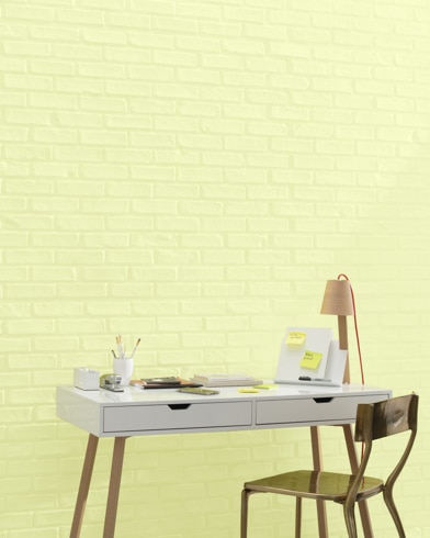 A white and copper writing desk and a bronze metal chair sit in front of a brick wall painted Lemon Twist.