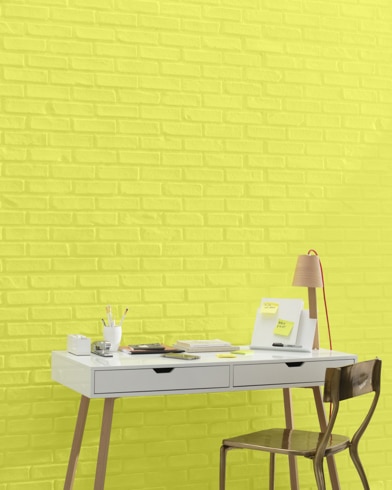 A white and copper writing desk and a bronze metal chair sit in front of a brick wall painted Limelight.