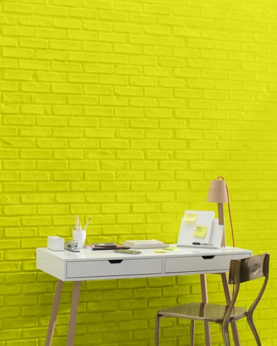 A white and copper writing desk and a bronze metal chair sit in front of a brick wall painted New Lime.