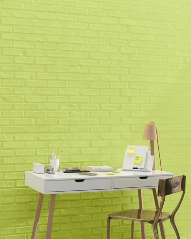 A white and copper writing desk and a bronze metal chair sit in front of a brick wall painted Pear Green.