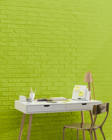 A white and copper writing desk and a bronze metal chair sit in front of a brick wall painted Tequila Lime.
