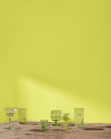 A variety of blue and green glass cocktail glasses sit on a table in front of a wall painted Granny Smith.