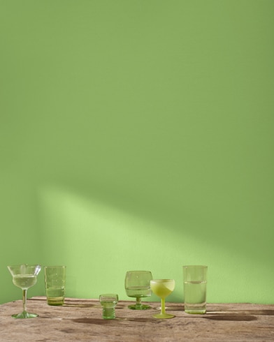 A variety of blue and green glass cocktail glasses sit on a table in front of a wall painted Green Thumb.