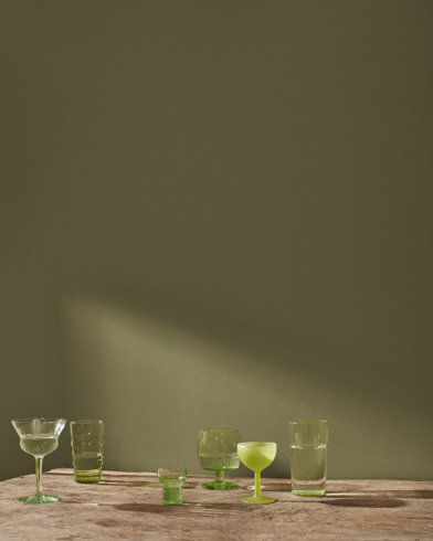 A variety of blue and green glass cocktail glasses sit on a table in front of a wall painted Jungle Canopy.