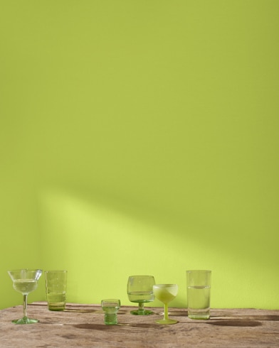 A variety of blue and green glass cocktail glasses sit on a table in front of a wall painted Limeade.