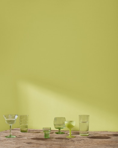 A variety of blue and green glass cocktail glasses sit on a table in front of a wall painted Martini Olive.