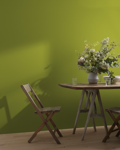 A wood folding chair sits next to a table topped with a greenery-filled vase in front of a Dark Celery-painted wall.