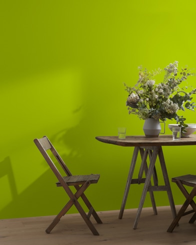 A wood folding chair sits next to a table topped with a greenery-filled vase in front of a Dark Lime-painted wall.