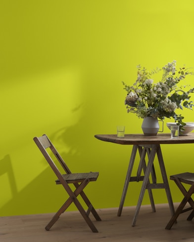 A wood folding chair sits next to a table topped with a greenery-filled vase in front of a Eccentric Lime-painted wall.