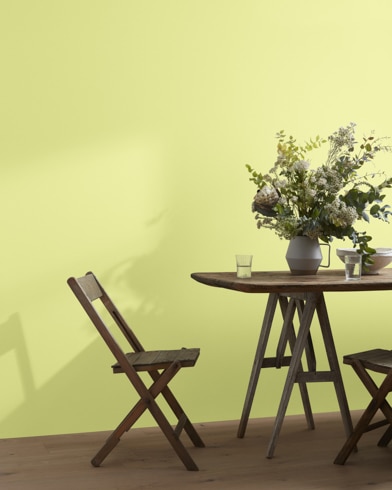 A wood folding chair sits next to a table topped with a greenery-filled vase in front of a Hibiscus-painted wall.