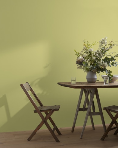 A wood folding chair sits next to a table topped with a greenery-filled vase in front of a Lilianna-painted wall.