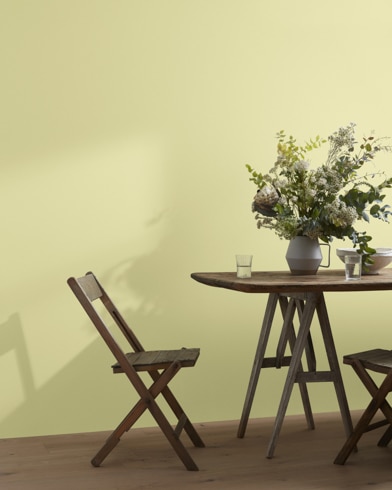 A wood folding chair sits next to a table topped with a greenery-filled vase in front of a Lime Sherbet-painted wall.