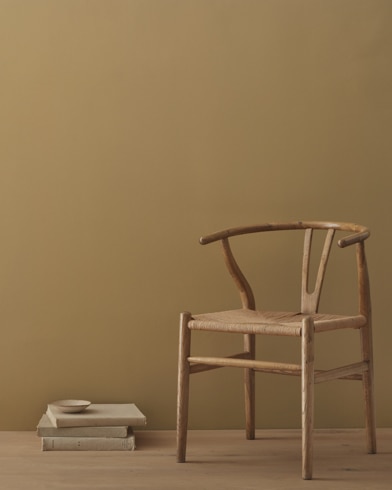 A modern wicker chair and a stack of books topped with a small bowl sit in front of a room painted Iced Coffee.