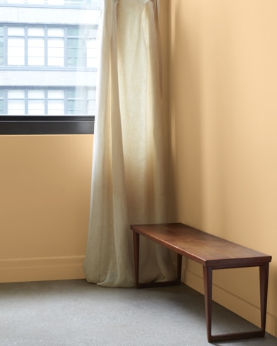 In a Classic Caramel-painted room with a wooden bench, a gauzy white curtain hangs next to an open window.