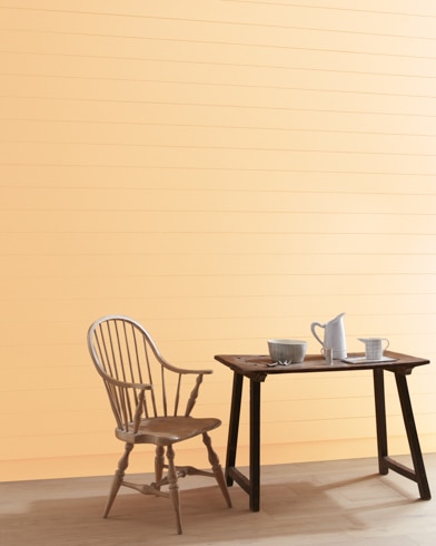 Una pared pintada de color Arena de Asbury con una silla con barrotes de madera y una jarra blanca, una taza y un bol sobre una mesa.