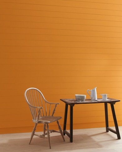 Una pared pintada de color Anaranjado del Otoño con una silla con barrotes de madera y una jarra blanca, una taza y un bol sobre una mesa.