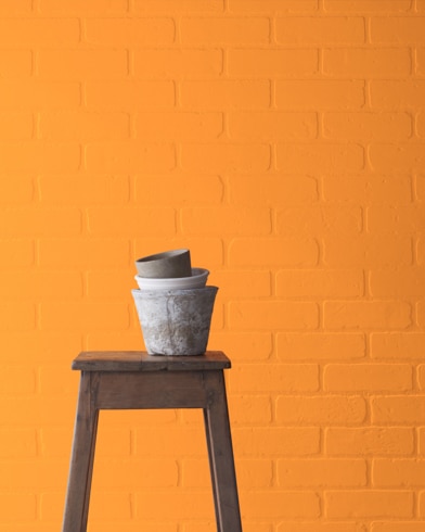 A Carrot Stick-painted brick wall behind a wooden stool with planting pots on top.