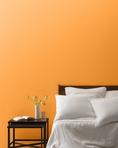 A Mango Punch-painted bedroom wall, bed with white linens, and a night stand.