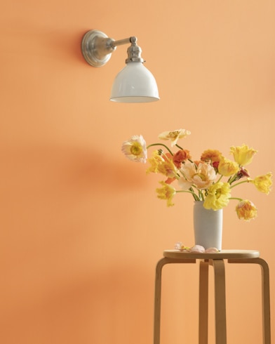 A Golden Dunes-painted wall with single sconce, and a wooden stool with flower-filled vase. 