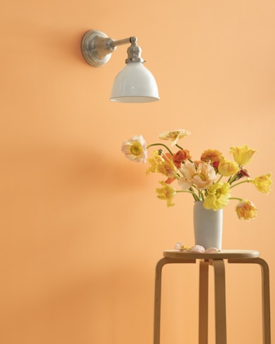 A Citrus Blossom-painted wall with single sconce, and a wooden stool with flower-filled vase. 