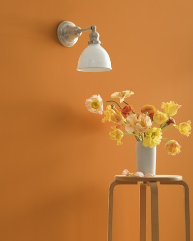 A Golden Dunes-painted wall with single sconce, and a wooden stool with flower-filled vase. 