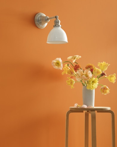 A Pumpkin Spice-painted wall with single sconce, and a wooden stool with flower-filled vase. 