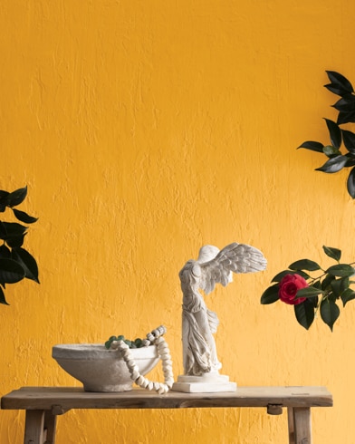 A statue and a bowl on a wooden bench in front of a textured wall painted in Yellow Marigold.