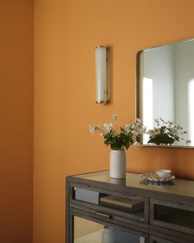 Una mesa marrón con un juego de té y un jarrón con flores debajo de un espejo colgado en una pared pintada de color Oro Veneciano.