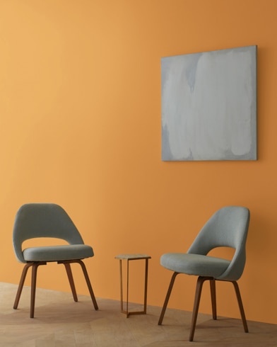 Two blue chairs and a table stand in front of a wall under an abstract painting hanging on a wall painted Amber.