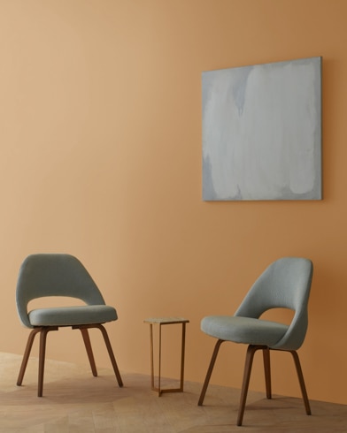 Two blue chairs and a table stand in front of a wall under an abstract painting hanging on a wall painted Etruscan.