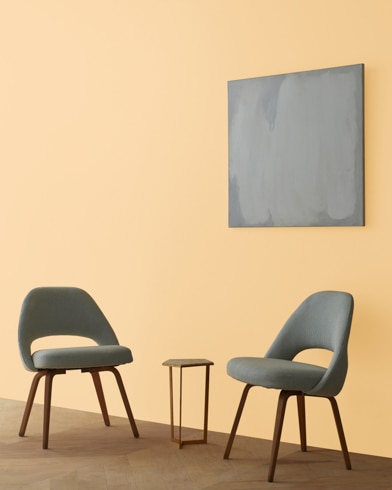 Two blue chairs and a table stand in front of a wall under an abstract painting hanging on a wall painted Glowing Apricot.