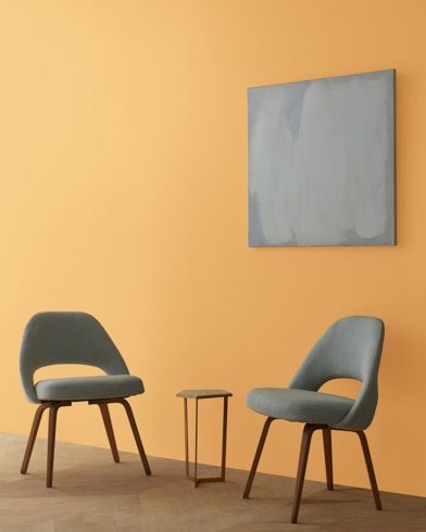 Two blue chairs and a table stand in front of a wall under an abstract painting hanging on a wall painted Old Gold.