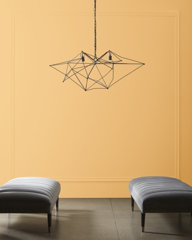 A modern, wiry chandelier hangs over two velvet ottomans in front of a wall painted Amber Waves.