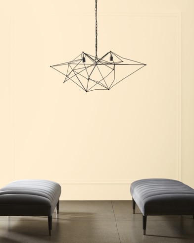 A modern, wiry chandelier hangs over two velvet ottomans in front of a wall painted Cream.