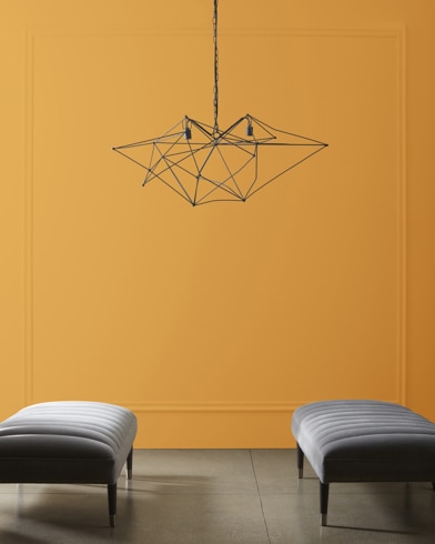 A modern, wiry chandelier hangs over two velvet ottomans in front of a wall painted Peanut Butter.