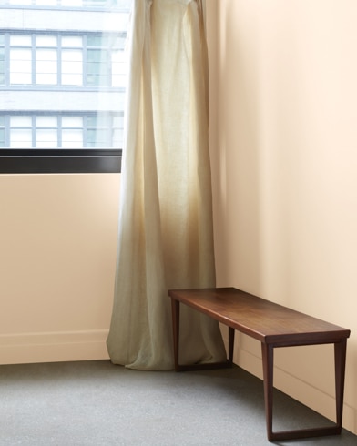 In a Painted Sands-painted room with a wooden bench, a gauzy white curtain hangs next to an open window.