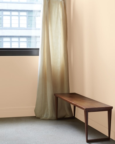 In a Powder Puff-painted room with a wooden bench, a gauzy white curtain hangs next to an open window.