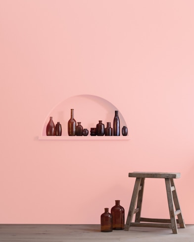 A wood stool and bottles in front of a Dawn Pink-painted wall with inset shelving holding a variety of empty bottles.