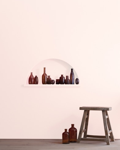 A wood stool and bottles in front of a Frosty Pink-painted wall with inset shelving holding a variety of empty bottles.