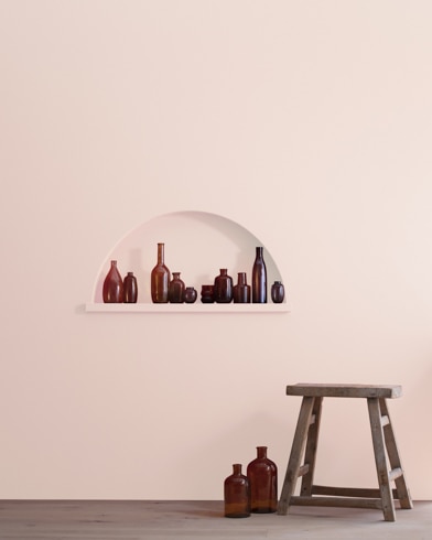 A wood stool and bottles in front of a Rose Accent-painted wall with inset shelving holding a variety of empty bottles.