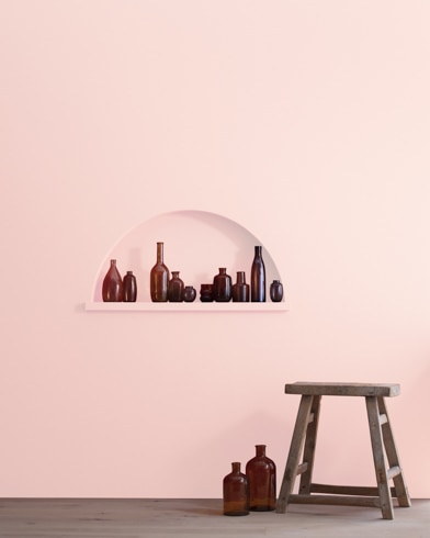 A wood stool and bottles in front of a Rose Petal-painted wall with inset shelving holding a variety of empty bottles.