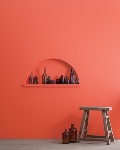 A wood stool and bottles in front of a Starburst Orange-painted wall with inset shelving holding a variety of empty bottles.