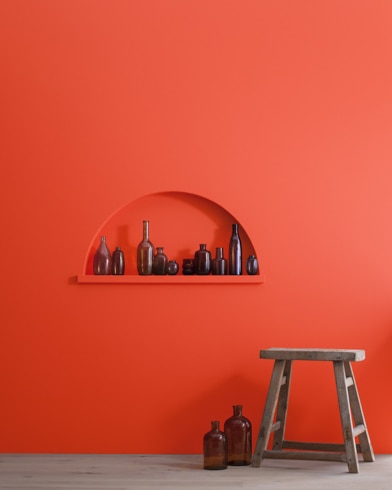 A wood stool and bottles in front of a Tomato Red-painted wall with inset shelving holding a variety of empty bottles.