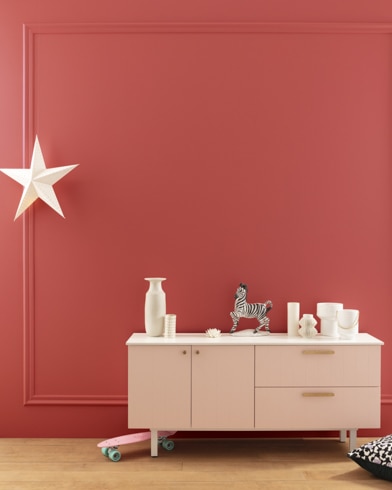 A modern white dresser with decor in front of a Rosy Apple-painted wall with square molding, and a dangling white star. 