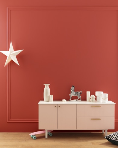 A modern white dresser with decor in front of a Sangria-painted wall with square molding, and a dangling white star. 